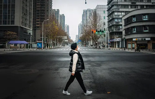 Foto colorida de um cenário atual, 2020, de uma mulher caminhando sozinha em uma rua completamente deserta da cidade.